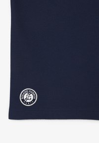 Mörkblå strukturerad sportskjorta i tyg med en cirkulär broderad logga med texten "Roland Garros" och "Paris" i fållen.