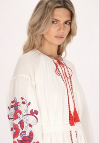 Weißes, besticktes Kleid mit roten Blumenmustern, langen Ärmeln und einer Bindung mit Quasten. Weicher Stoff und luftige Passform.