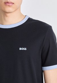 Svart ribbestrikket t-skjorte med lyseblå krage og mansjettkanter. Har en liten hvit "BOSS" logo på brystet. Glatt tekstur.
