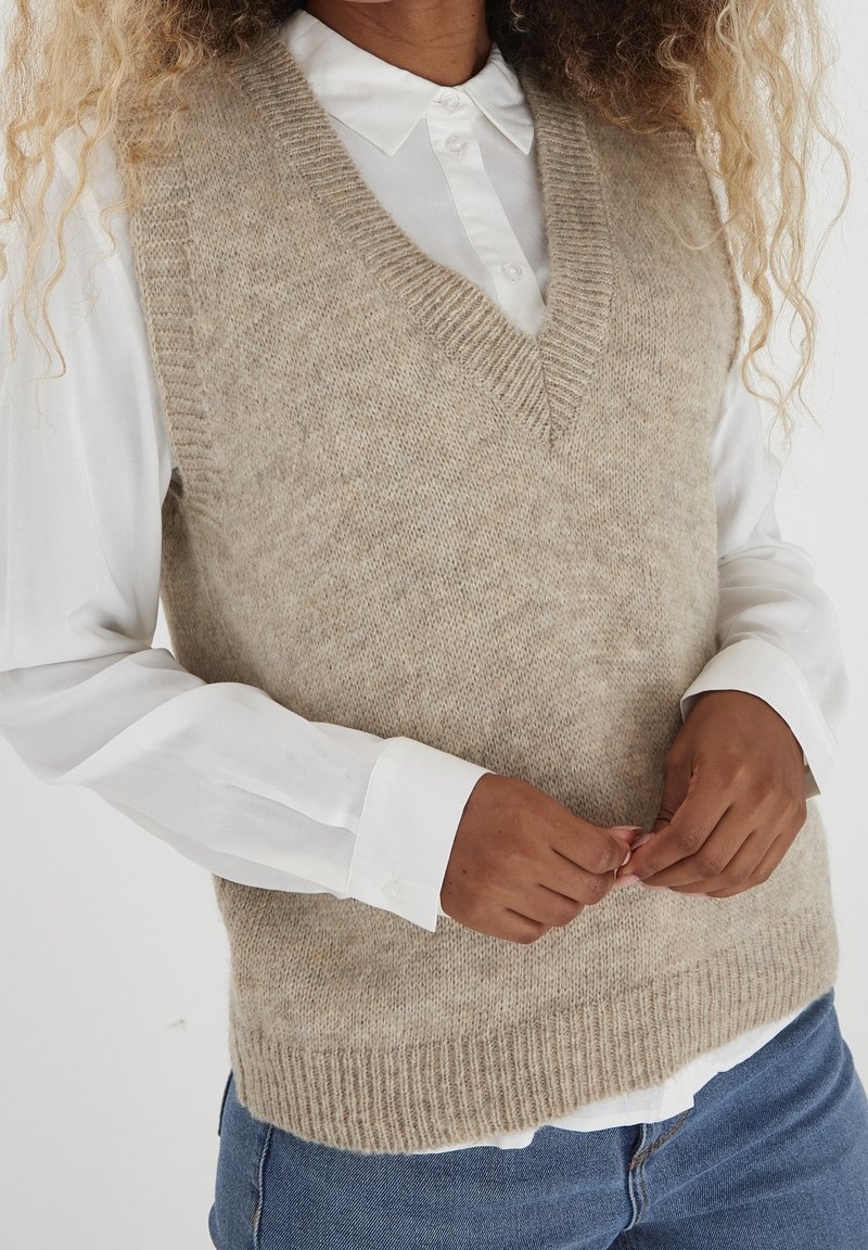 Personne portant un gilet beige en maille sans manches sur une chemise blanche à col et un jean bleu, mains doucement croisées au niveau de la taille.