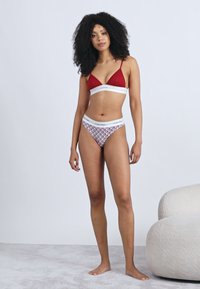 Červená trojuholníková podprsenka s bielym spodným pásom, v páre s bielymi a vzorovanými bikinovými nohavičkami s pásom s logom značky. Hladká textúra.