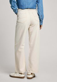 Jeans de pierna ancha de color claro con sutiles rayas verticales, cuatro bolsillos y costuras traseras visibles. Llevados con una camisa de mezclilla azul y zapatillas.