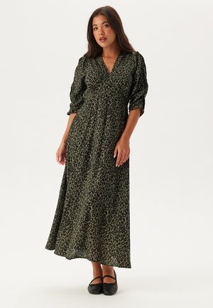Maxiklänning i mörkgrön med svart leopardtryck, med V-ringning, pösiga ärmar och en luftig textur, tillsammans med svarta platta skor.