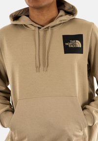 Felpa beige realizzata in tessuto morbido, dotata di tasca frontale, cappuccio regolabile con cordino e un logo quadrato nero sul petto.