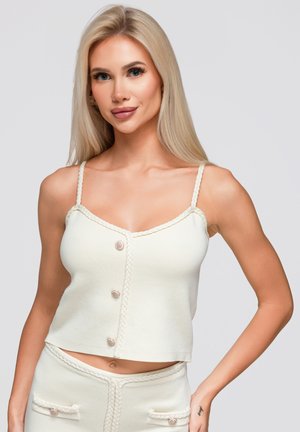 Femme blonde portant un crop top en maille blanc avec des boutons et un pantalon taille haute assorti, debout devant un fond uni.