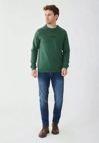 Felpa verde con scollatura rotonda e una piccola stampa nera sul petto, abbinata a jeans blu e stivali marroni con lacci.