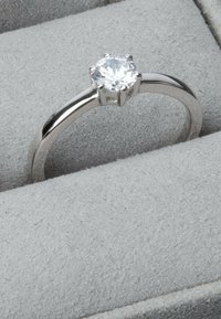 Silver ring med en rund, klar diamant som är inställd i en sexklorad fäste, vilande på grå mjuk padding i en visningslåda.