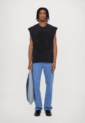 Black sleeveless top with large textured graphic, paired with light blue jeans and black shoes, holding a gray jacket.