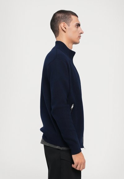 Chaqueta de punto con cremallera en color navy y cuello alto, con un bolsillo lateral y un diseño ajustado. Textura suave y corte casual.