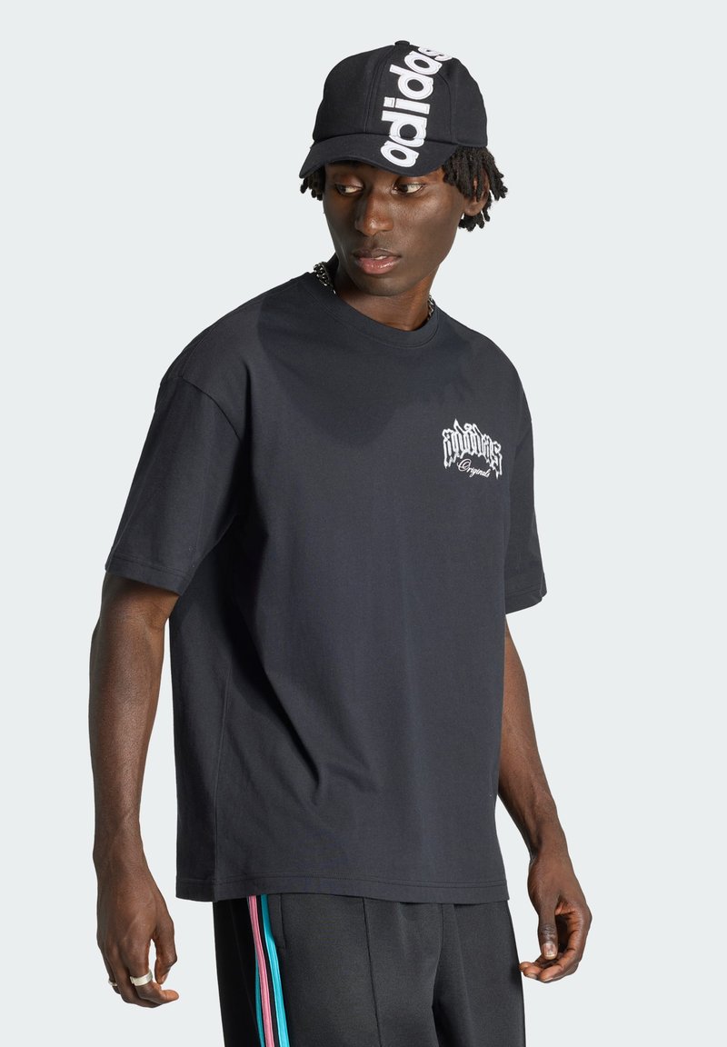 Joven con una gorra negra de Adidas, camiseta negra con estampado gráfico y pantalones negros con franjas laterales azules y rosas, mirando hacia abajo a su izquierda.