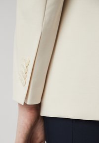 Manga de un blazer beige claro, que presenta una textura suave y tres botones redondos con un acabado brillante cerca del puño, diseñado para un ajuste ceñido.