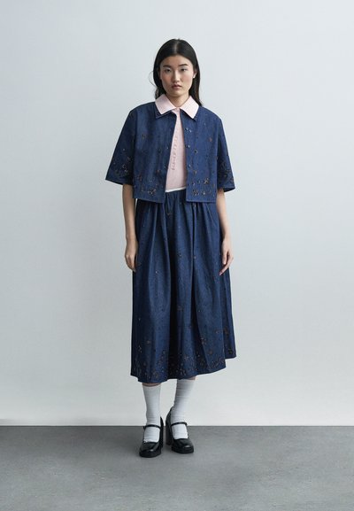 Femme portant une veste et une jupe brodées bleu marine, un haut rose à col, des chaussettes blanches et des escarpins noirs de type Mary Jane, debout devant un fond uni.