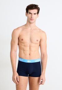 Tamsiai mėlyni boxer tipo apatiniai su šviesiai mėlynu Lacoste logotipo juosmeniu. Medžiaga atrodo lygi ir elastinga, su prigludusiu dizainu ir jokių matomų raštų.