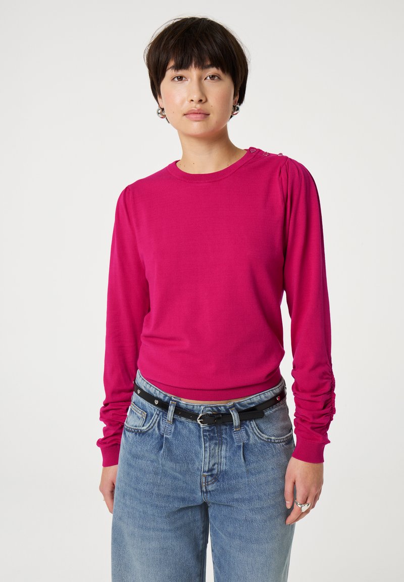 Getailleerde langemouwen top in felroze breiwerk, met geplooide details op de schouders en een ronde halslijn, gecombineerd met lichtblauwe high-waisted jeans.