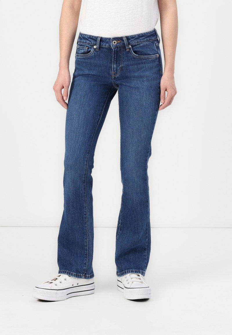 Flared dark blue jeans made of denim with a mid-rise waist, five pockets, subtle fading, and raw edge hem, paired with white sneakers.