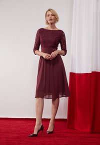 Robe mauve avec un corsage en dentelle et des manches trois-quarts, accompagnée d'une jupe plissée fluide. Associée à des talons noirs pointus sur un tapis rouge.