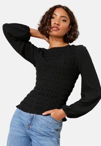 Kobieta z krótkimi, kręconymi włosami, nosząca złote kolczyki w kształcie kół, strukturalny czarny top z długim rękawem i jasnoniebieskie dżinsy, pozująca z jedną ręką na szyi.