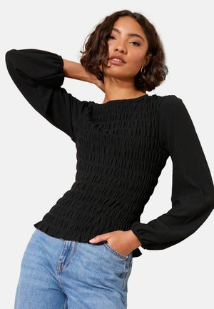 Frau mit kurzen lockigen Haaren, die goldene Creolen, ein strukturiertes schwarzes Langarmoberteil und hellblaue Jeans trägt und mit einer Hand am Hals posiert.