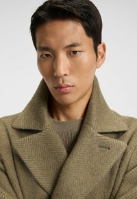 Houndstooth-patterned wool coat in olive green with a wide collar and textured fabric. The model wears a ribbed brown sweater underneath.