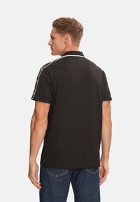 Camisa polo negra con cuello blanco y rayas laterales, hecha de tela texturizada. La vista trasera muestra mangas cortas y un sutil detalle del logo.