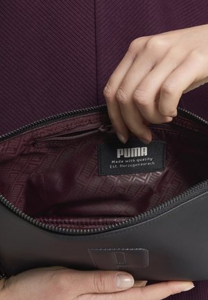 Bolsa de cuero negro con interior burdeos, con cierre de cremallera y una etiqueta que muestra el logo de Puma y los detalles de la marca.