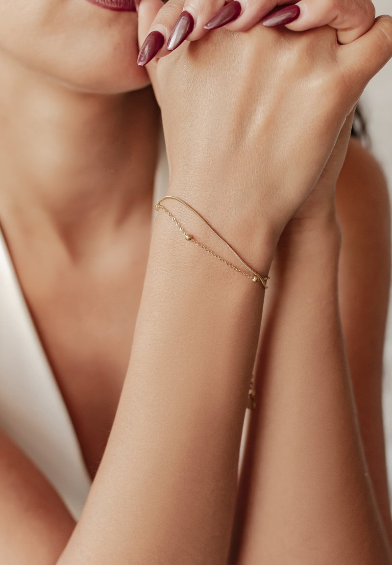 Pulsera de cadena de oro con un diseño sencillo y una pequeña cuenta redonda, llevada en una mano con un tono de piel natural y uñas largas y pulidas.