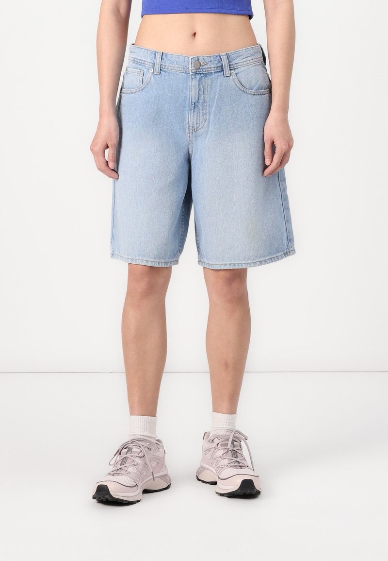 Shorts en denim bleu clair à coupe droite, avec poches avant et fermeture à boutons. Associés à des chaussures de sport pâles et des chaussettes blanches.