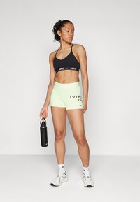 Sujetador deportivo negro con el logo de Nike, pantalones cortos amarillos neón con un logo en contraste, sosteniendo una botella de agua negra y usando zapatillas deportivas blancas.