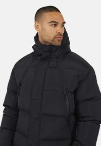 Doudoune noire avec un col montant, des poches zippées et un design matelassé. Le matériau semble lisse et léger.