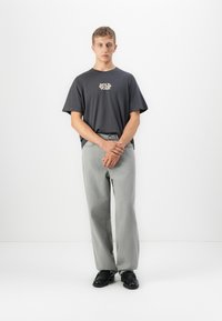 Young man standing, wearing a dark gray "Wild Soul" t-shirt, light gray jeans, and black shoes against a plain white background.