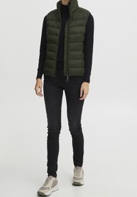 Quiltad grön pufferväst med hög krage, dragkedja framtill, bärs över en svart långärmad topp, kombineras med svarta tight jeans och ljusa sneakers.