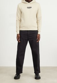 Felpa con cappuccio beige con logo sul petto, abbinata a pantaloni neri a vestibilità larga e stivali neri chunky, in piedi su uno sfondo neutro.