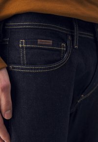 Tmavě modré džíny z denimu s kontrastním žlutým prošíváním, vybavené malou koženou nášivkou s logem na zadním kapse a standardními očky na pásek.