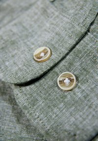 Tela de lino verde con un acabado texturizado, que presenta dos botones redondos de color claro asegurados con hilo blanco en un detalle de solapa.