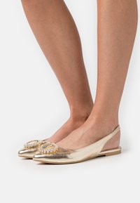 Gold metallic flats with a pointed toe, featuring a decorative gem-encrusted accent at the front and a slingback design. Smooth texture.
