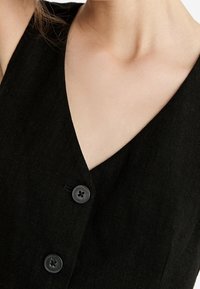 Czarna lniana sukienka z głębokim dekoltem w kształcie V, dwiema czarnymi guzikami i teksturowanym materiałem, podkreślająca prosty, funkcjonalny design.