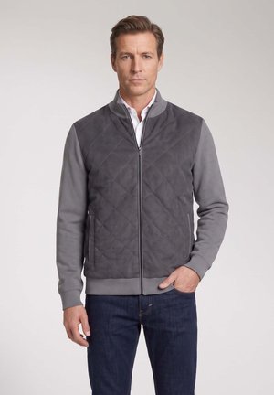 Mann mit kurzen braunen Haaren, der eine graue gesteppte Reißverschlussjacke über einem weißen Hemd und dunkelblauen Jeans trägt, steht vor einem schlichten Hintergrund.