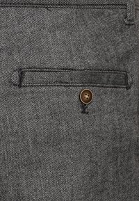 Šedá tkanina v rybí kost s texturovaným povrchem, vybavená klopovým kapsou s tmavým knoflíkem. Jemné a pečlivé švy podél okrajů.