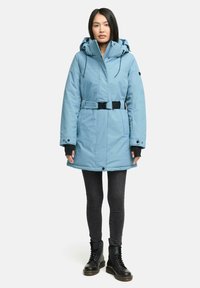 Ljusblå isolerad parkas med huva och dragsko vid midjan, med svarta detaljer, blixtlås och tryckknappar, bärs över svarta skinny jeans och stövlar.