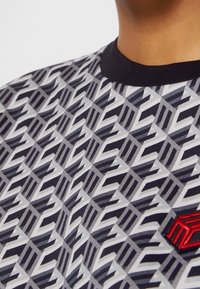 Zbliżenie osoby w czarnej koszuli z kołnierzykiem, na której znajduje się szaro-biała geometryczna wzór oraz mała czerwona haftowana emblemat.