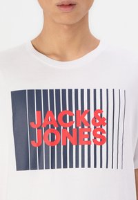 Tricou alb cu un design grafic albastru și roșu, având textul "JACK & JONES" și dungi verticale în fundal.