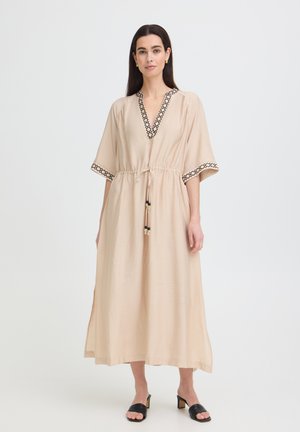 Mujer con vestido largo beige con ribete geométrico negro en las mangas y el escote, de pie con sandalias negras de tacón sobre fondo blanco.