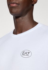 Bela bomba z vlečnim ovratnikom iz bombaža z črnim okroglim logotipom, ki na levi strani nosi napis "EA7 EMPORIO ARMANI". Gladka tekstura.