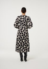 Maxi-jurk van lichtgewicht stof met een zwart hartjespatroon op een beige achtergrond. Lange pofmouwen en een ceintuur in de taille. Zwarte kniehoge laarzen.