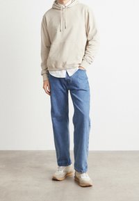 Beige hoodie med framficka, bärs över en vit skjorta, kombinerad med ljusblå denimjeans och beige sneakers med grå detaljer.