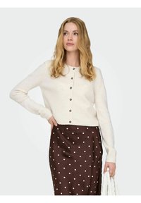 Cardigan lavorato a maglia color crema con scollo rotondo e bottoni frontali, abbinato a una gonna a pois marroni. Texture liscia e design attillato.