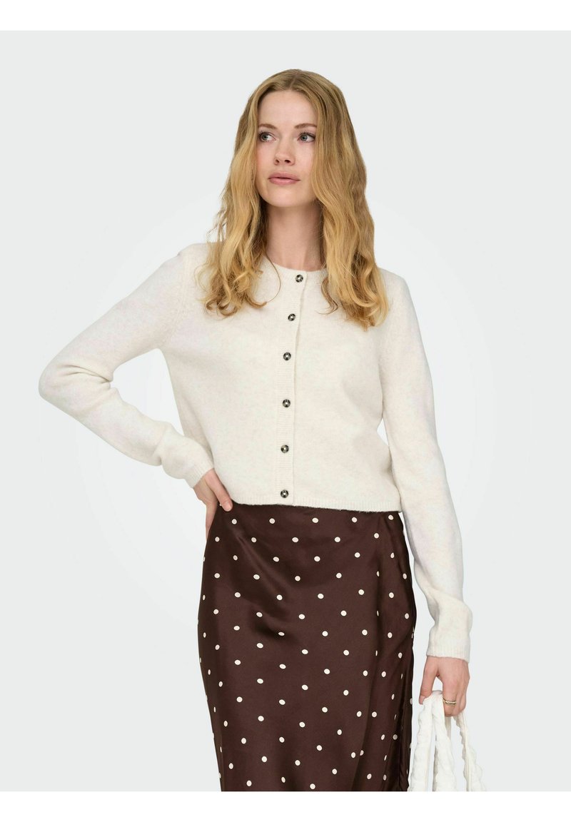 Cardigan lavorato a maglia color crema con scollo rotondo e bottoni frontali, abbinato a una gonna a pois marroni. Texture liscia e design attillato.