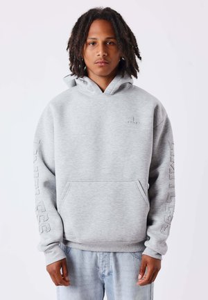 Young person with medium-length dreadlocks wearing a light gray hoodie with text on sleeves and light blue jeans standing against a plain background.