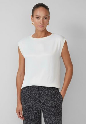 White, short-sleeve top with a round neckline, made from smooth fabric. Worn with textured dark patterned pants. Simple and clean design.