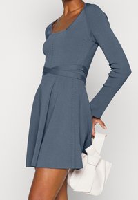 Modré šaty s dlouhým rukávem a čtvercovým výstřihem, s texturovaným pleteným vzorem, střihaným živůtkem, rozšířenou sukní a páskem ze stejné látky. Bílá kabelka.